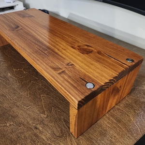 May include: A wooden monitor stand with a natural wood finish. The stand has a simple design with a rectangular top and a raised base. The stand is made of solid wood and has a smooth, polished surface.