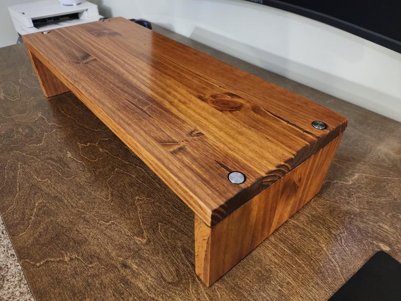 May include: A wooden monitor stand with a natural wood finish. The stand has a simple design with a rectangular top and a raised base. The stand is made of solid wood and has a smooth, polished surface.