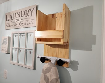 May include: A wooden wall-mounted shelf unit with a sign that reads "Laundry Room Self service, Open 24/7". The shelf has a small shelf on top and a larger shelf below. There is a white iron with a gray cover hanging on a hook below the shelf.