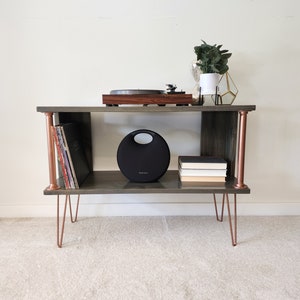 May include: A dark gray wooden record player stand with copper pipe accents and hairpin legs. The stand has two shelves, one for a record player and the other for records and books.