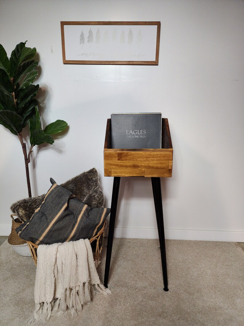 May include: A wooden record stand with a dark stain finish, featuring a black metal leg base. The stand holds a record album with the text "EAGLES THE LONG RUN". A framed art piece with a forest scene hangs on the white wall.