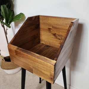 May include: A wooden storage bin with a warm brown finish, set on black metal hairpin legs. The bin has a slanted front and is constructed from wood planks. The design is simple and modern, suitable for various storage needs.