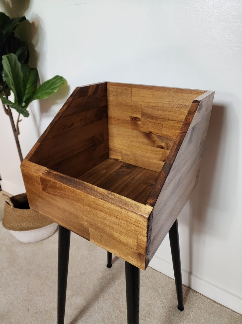 May include: A wooden storage bin with a warm brown finish, set on black metal hairpin legs. The bin has a slanted front and is constructed from wood planks. The design is simple and modern, suitable for various storage needs.