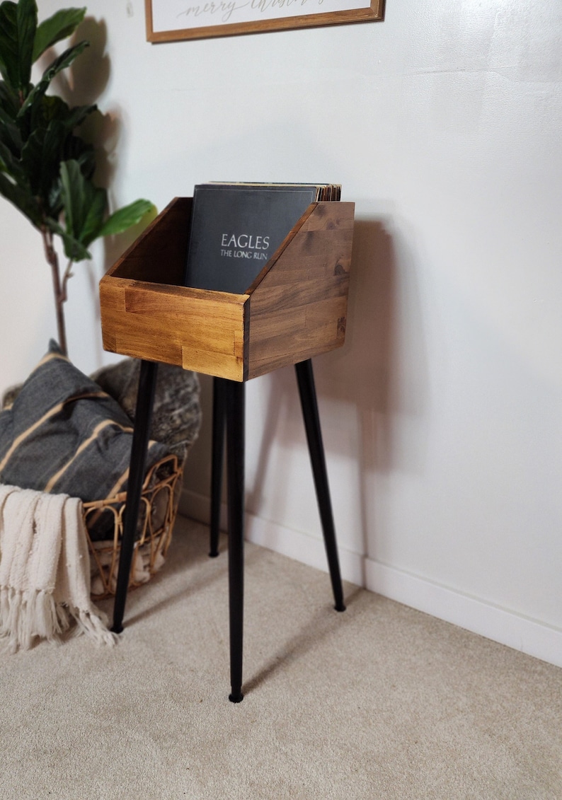 May include: Wooden record storage stand with a dark stain finish. The stand has a rectangular bin with angled sides, supported by black metal legs. The bin holds vinyl records, with the album "Eagles The Long Run" visible.
