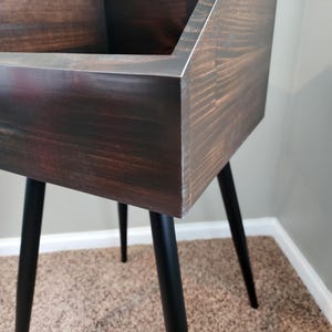May include: A dark stained wooden storage unit with a rectangular top and black metal legs. The unit has a mid-century modern design and is suitable for various storage needs. The legs are angled and provide stability.