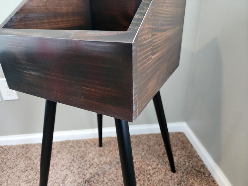 May include: A dark stained wooden storage unit with a rectangular top and black metal legs. The unit has a mid-century modern design and is suitable for various storage needs. The legs are angled and provide stability.