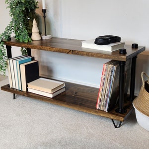 May include: A two-tiered industrial style shelf unit with black metal pipes and a dark wood finish. The shelf is decorated with books, records, and a small wooden ornament.