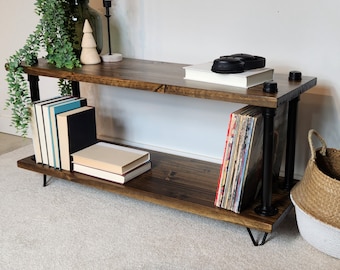 May include: A two-tiered industrial style shelf unit with black metal pipes and a dark wood finish. The shelf is decorated with books, records, and a small wooden ornament.