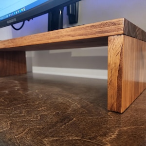 May include: A wooden monitor stand with a natural wood finish. The stand has a rectangular top and two vertical supports. The stand is made of solid wood and has a simple, modern design.