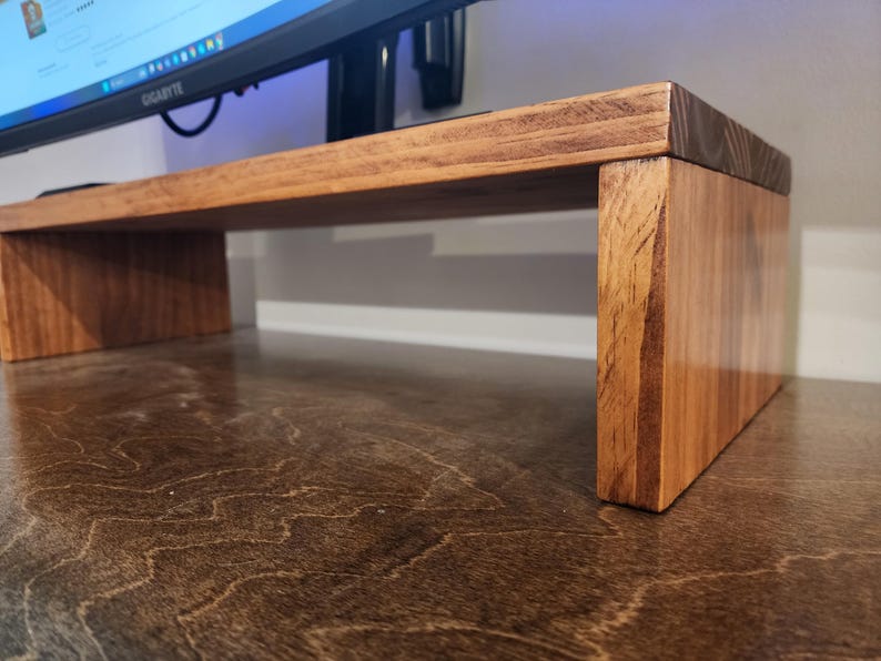 May include: A wooden monitor stand with a natural wood finish. The stand has a rectangular top and two vertical supports. The stand is made of solid wood and has a simple, modern design.