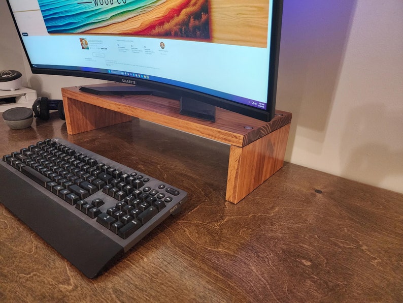 May include: A wooden monitor stand with a curved monitor on top. A black keyboard is in the foreground. The stand is made of natural wood with a light brown finish.