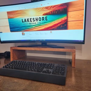 May include: A curved monitor with a wooden stand sits on a wooden desk. The monitor is displaying a website for "Lakeshore Wood Co." with a colorful image of a beach scene. A black keyboard is in the foreground.