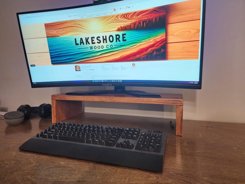 May include: A curved monitor with a wooden stand sits on a wooden desk. The monitor is displaying a website for "Lakeshore Wood Co." with a colorful image of a beach scene. A black keyboard is in the foreground.