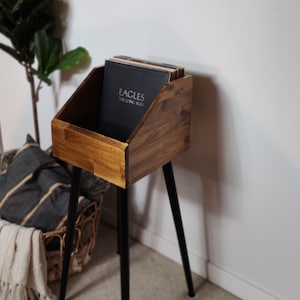 May include: A wooden record stand with a dark stain finish, holding vinyl records. The stand has a rectangular bin shape and sits on three black metal legs. A framed sign with a Christmas theme hangs on the wall.
