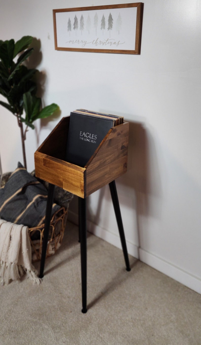 May include: A wooden record stand with a dark stain finish, holding vinyl records. The stand has a rectangular bin shape and sits on three black metal legs. A framed sign with a Christmas theme hangs on the wall.