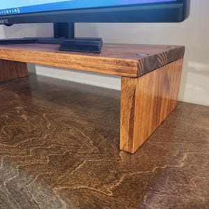 May include: A wooden monitor stand with a natural wood finish. The stand is made of solid wood and has a simple, modern design. It is perfect for raising a monitor to a more ergonomic height.