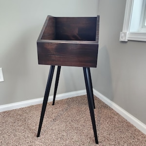 May include: A dark brown wooden storage stand with a rectangular box and angled black legs. The stand has a minimalist design and is suitable for various storage needs. The stand is placed on a carpeted floor.