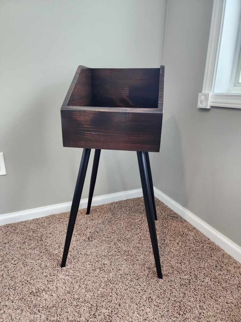 May include: A dark brown wooden storage stand with a rectangular box and angled black legs. The stand has a minimalist design and is suitable for various storage needs. The stand is placed on a carpeted floor.