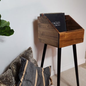 May include: A wooden record stand with black metal legs holds vinyl records. A woven basket with pillows and a blanket sits below. A framed sign above reads "merry christmas".