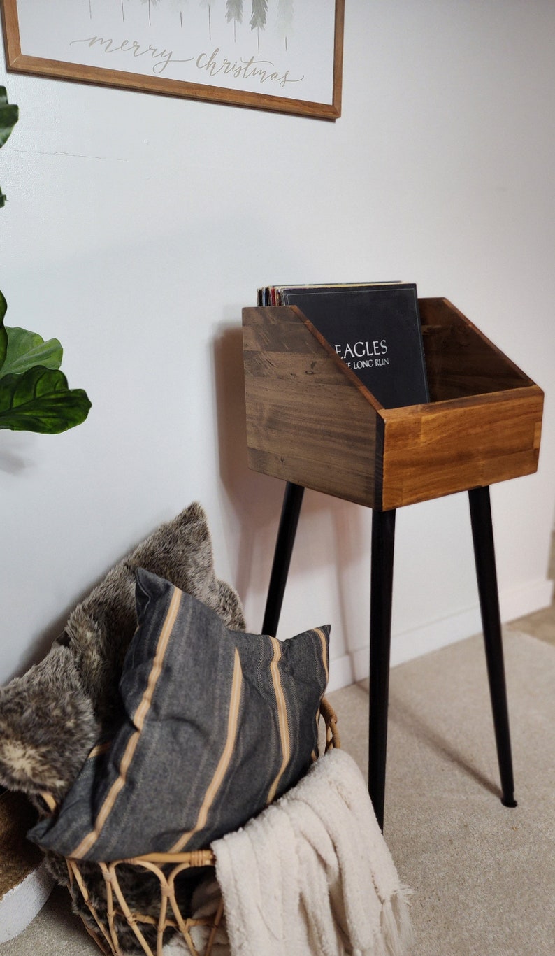 May include: A wooden record stand with black metal legs holds vinyl records. A woven basket with pillows and a blanket sits below. A framed sign above reads "merry christmas".