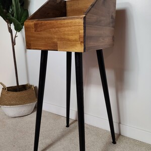 May include: A wooden record stand with a rectangular storage compartment and angled sides. The stand is made of light and dark brown wood and has four black, tapered legs. The stand is designed to hold vinyl records.