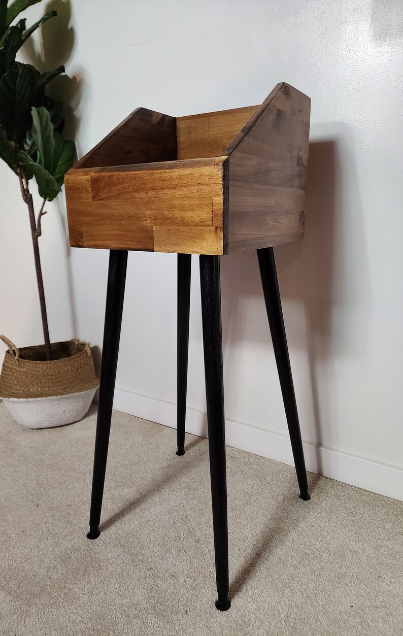 May include: A wooden record stand with a rectangular storage compartment and angled sides. The stand is made of light and dark brown wood and has four black, tapered legs. The stand is designed to hold vinyl records.