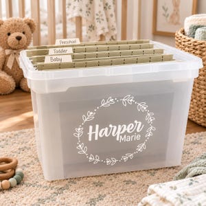 May include: A clear plastic storage bin featuring a white vinyl decal of a floral wreath surrounding the name Harper Marie. Inside the bin are several light green hanging file folders with tabs labeled Baby, Toddler, and Preschool.