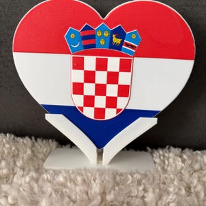 Könnte beinhalten: Eine herzförmige Plakette mit der kroatischen Flagge, die aus horizontalen roten, weißen und blauen Streifen mit dem nationalen Wappen in der Mitte besteht. Die Plakette wird von einem weißen, V-förmigen Kunststoffständer gehalten.