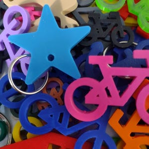 Op de afbeelding: Een verzameling kleurrijke sleutelhangers en bedels in verschillende vormen, waaronder sterren, fietsen en vredestekens. De sleutelhangers zijn in tinten blauw, roze, groen, oranje en paars. Een blauwe ster sleutelhanger staat op de voorgrond.