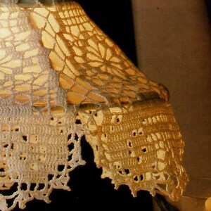 May include: A close-up of a white crocheted lampshade with a delicate floral pattern. The lampshade is lit from within, casting a warm glow.