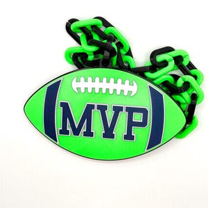 Könnte beinhalten: Eine grüne, Football-förmige Dekoration mit dem Wort "MVP" in Marineblau. Der Football hat weiße Schnürsenkel und ist marineblau umrandet. Eine schwarze und grüne Kette ist oben befestigt.