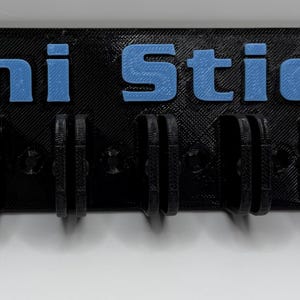 May include: A black holder with the words "Mini Sticks" in blue. The holder has multiple slots for holding small items. The holder is made of a matte black material and is mounted on a white surface.