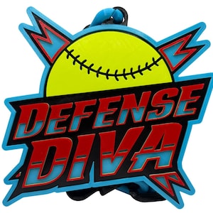 May include: A softball-themed pendant with the words "DEFENSE DIVA" in red, outlined in black, and set against a light blue background. The pendant features a yellow softball with black stitching and a blue loop for hanging.