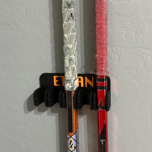 May include: Two hockey sticks, one orange and black, the other red and black, are held in a black holder mounted on a gray wall. The orange stick has the name "Ethan" on the holder.