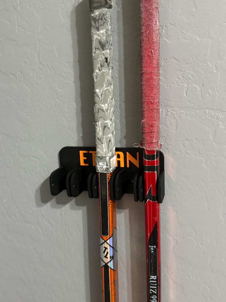 May include: Two hockey sticks, one orange and black, the other red and black, are held in a black holder mounted on a gray wall. The orange stick has the name "Ethan" on the holder.