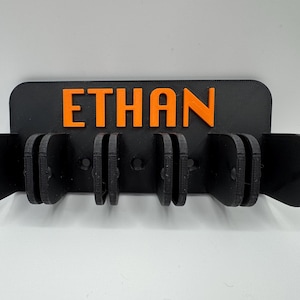 May include: A black wall-mounted holder with the name "ETHAN" in orange letters. The holder has multiple slots for organizing items. The design is simple and modern, suitable for various spaces.