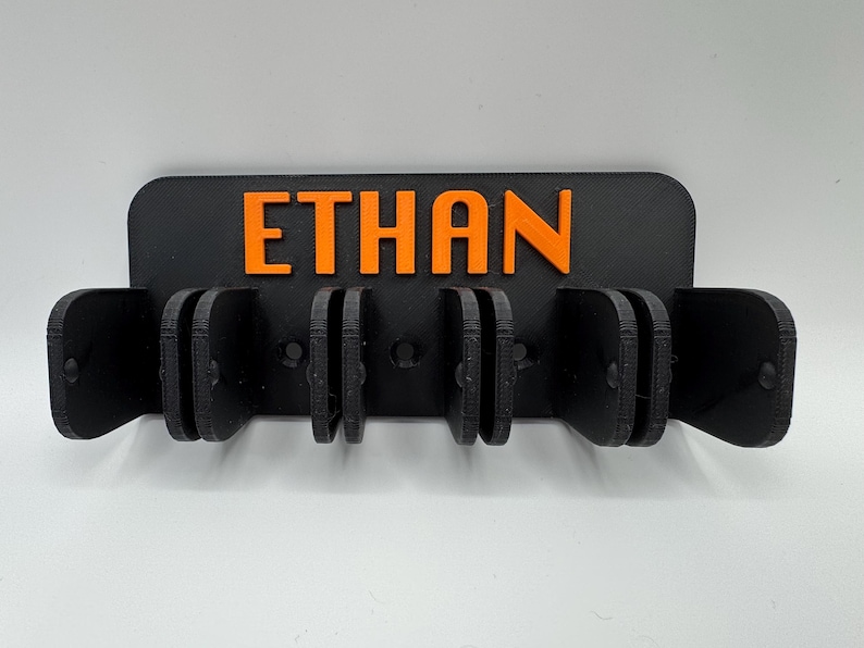 May include: A black wall-mounted holder with the name "ETHAN" in orange letters. The holder has multiple slots for organizing items. The design is simple and modern, suitable for various spaces.