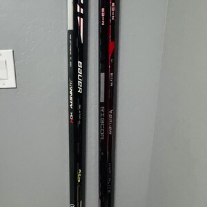 May include: Three hockey sticks leaning against a gray wall. One stick has a design of the American flag. The sticks are black and white with tape on the handles. The brands Bauer and Ribcor are visible.