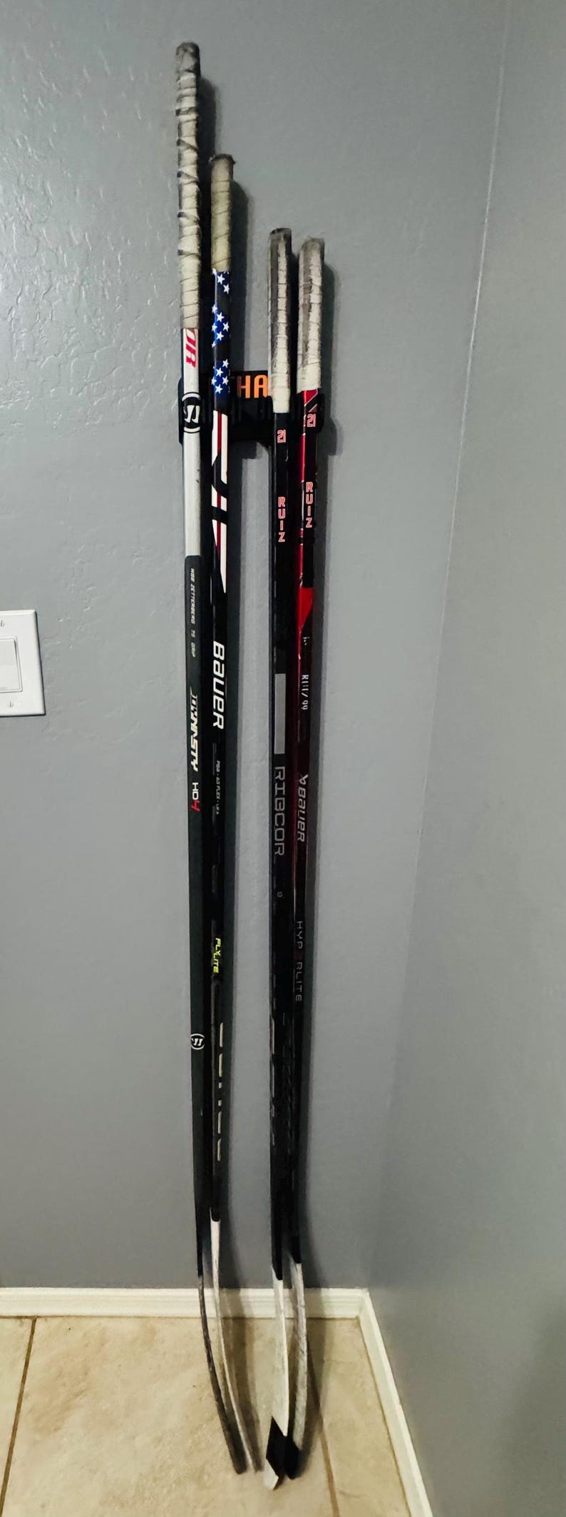 May include: Three hockey sticks leaning against a gray wall. One stick has a design of the American flag. The sticks are black and white with tape on the handles. The brands Bauer and Ribcor are visible.