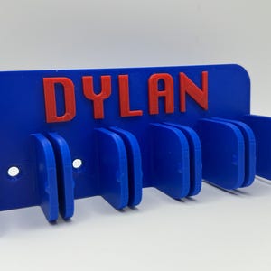 May include: A blue wall-mounted organizer with the name "DYLAN" in red letters. The organizer has multiple slots for holding items and two holes for mounting. The design is 3D printed.