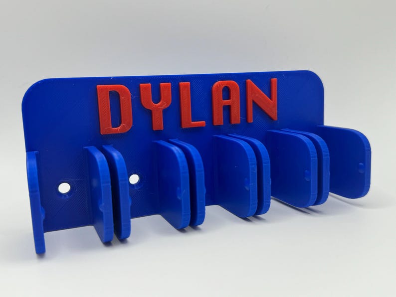 May include: A blue wall-mounted organizer with the name "DYLAN" in red letters. The organizer has multiple slots for holding items and two holes for mounting. The design is 3D printed.