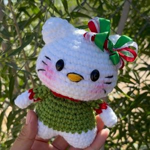 May include: A white crocheted cat figure with black oval eyes, a yellow nose, and pink stitched cheeks. The figure wears a green sweater with red and white trim on the sleeves and a festive red, white, and green ribbon bow on its head.