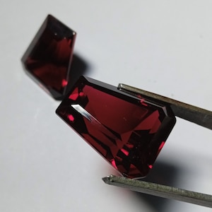 May include: Two red gemstone cabochons, one held by tweezers. The gemstones are cut in a rectangular shape with bevelled edges.