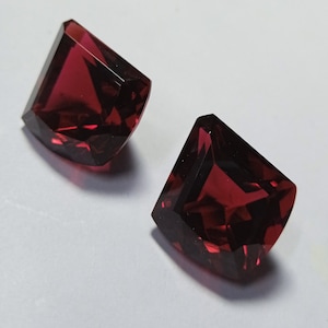 May include: Two faceted, deep red gemstones with a geometric cut. The gems are displayed on a white surface, showcasing their clarity and rich colour. The cut of the stones creates multiple reflective surfaces.