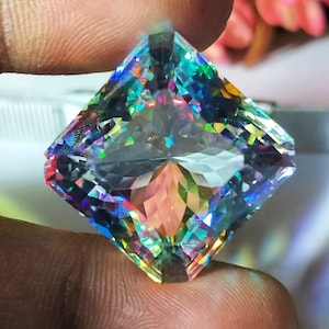 Può includere: Una gemma trasparente e sfaccettata con forma quadrata e colori arcobaleno iridescenti. La gemma è tenuta in una mano.