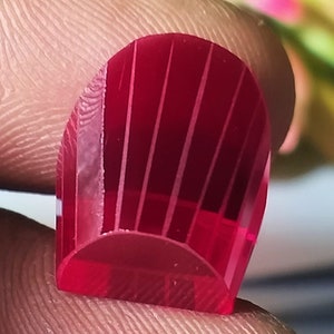 May include: A deep red, faceted gemstone with a rounded, slightly pointed top. The gemstone has a striated pattern on its surface.