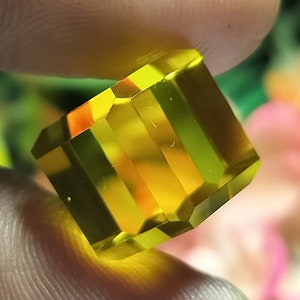 May include: A faceted, rectangular gemstone with a vibrant yellow hue. The gem is held between fingers, showcasing its polished surfaces and internal clarity. The cut reveals multiple facets, reflecting light and creating a brilliant appearance. The gemstone's color ranges from a bright yellow to a deeper amber.