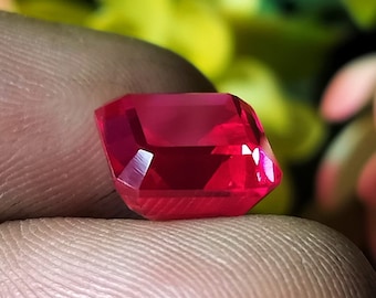 May include: A deep red gemstone with a rectangular emerald cut. The gemstone is faceted and has a high polish.