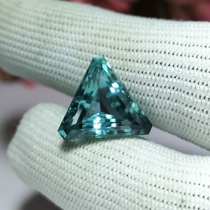 Könnte beinhalten: Ein dreieckiger, facettierter Edelstein mit einer blaugrünen Farbe. Der Edelstein ist vor einem weißen Hintergrund platziert.