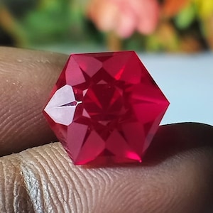 Może przedstawiać: Fasetowany, sześciokątny kamień szlachetny o głębokim, czerwonym odcieniu. Szlif kamienia tworzy liczne powierzchnie odbijające światło, zwiększając jego blask. Kamień trzymany jest między palcami, z rozmytym tłem w kolorze zielonym i pomarańczowym.